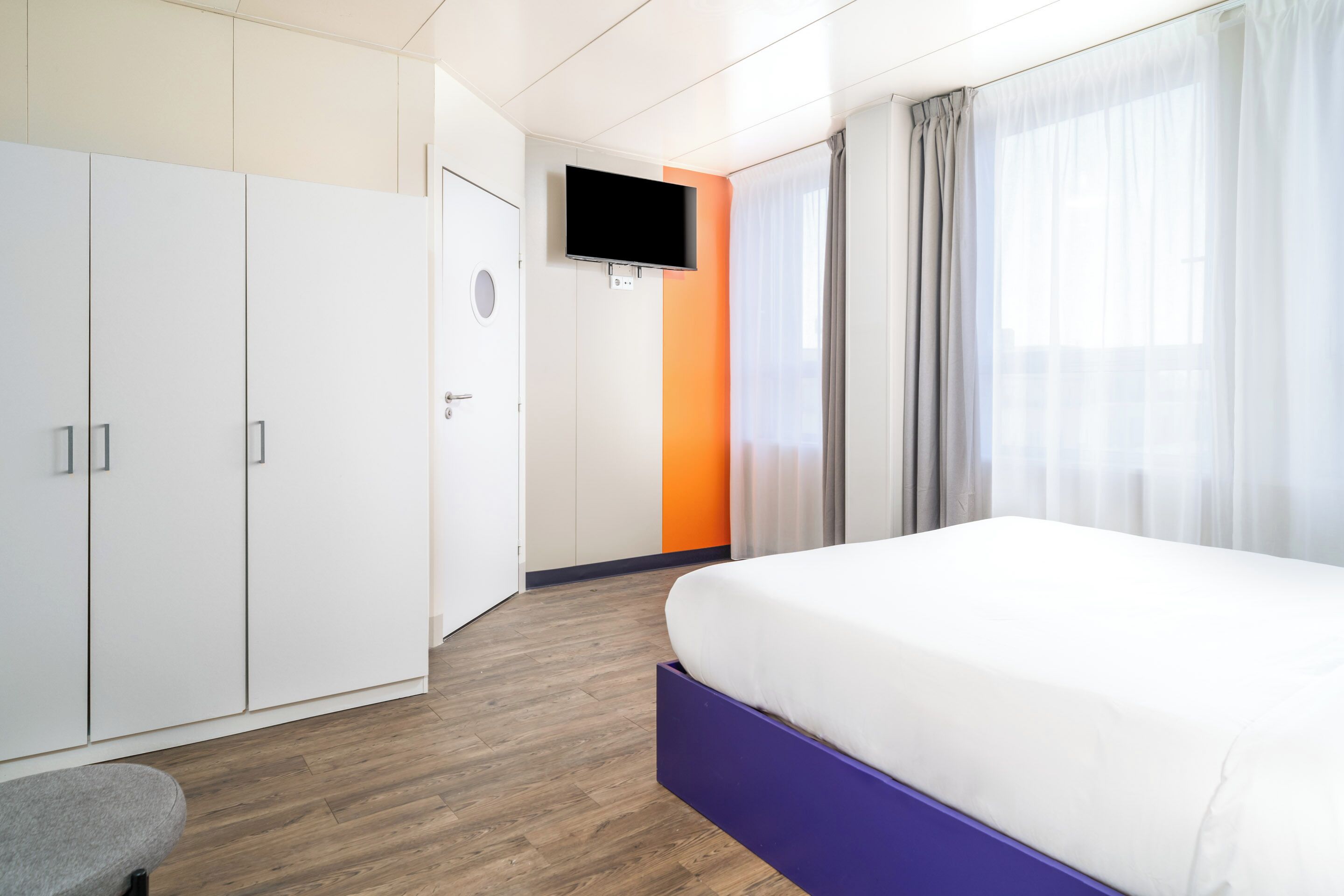 A modern hotel room with a large bed, wardrobe, and a flat-screen TV mounted on the wall.