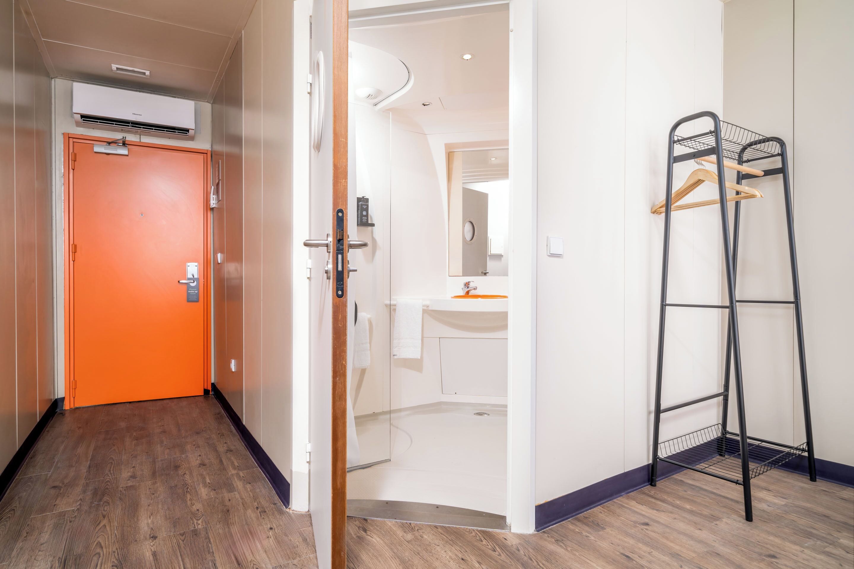 A hotel room with a wooden floor, a bathroom with a sink and mirror, and a black metal rack with wooden hangers.