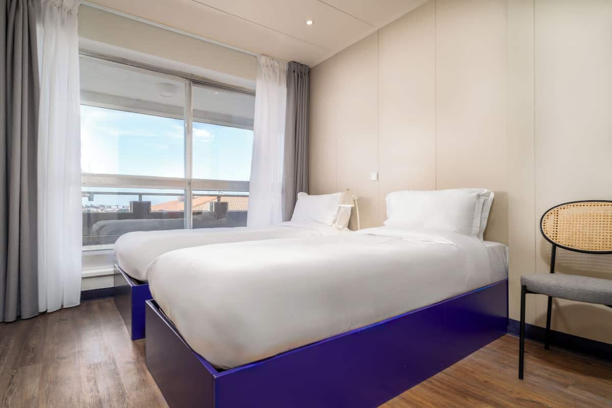 A hotel room with two beds, a large window, a chair, and a view of the city.