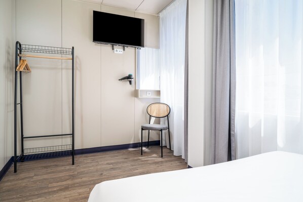 Habitación de hotel con suelo de madera, una cama, televisor montado en la pared, una silla y un perchero.