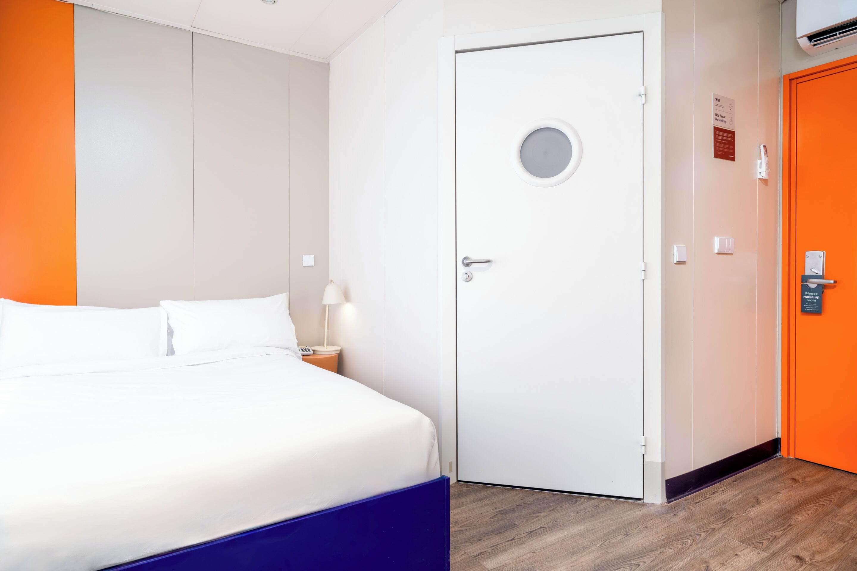 Et hotelværelse med en seng, et natbord, en hvid dør og en orange væg.