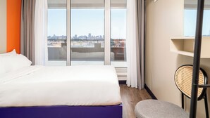 A hotel room with a bed, a chair, a TV, and a view of a cityscape through large windows. - STAYSOME Porto Herculano (Porto)