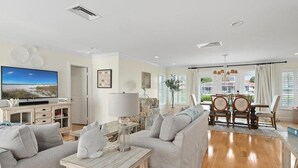 Uma sala de estar espaçosa com uma televisão de tela grande, sofás cinzas, uma mesa de centro de madeira e uma área de jantar com um candelabro.