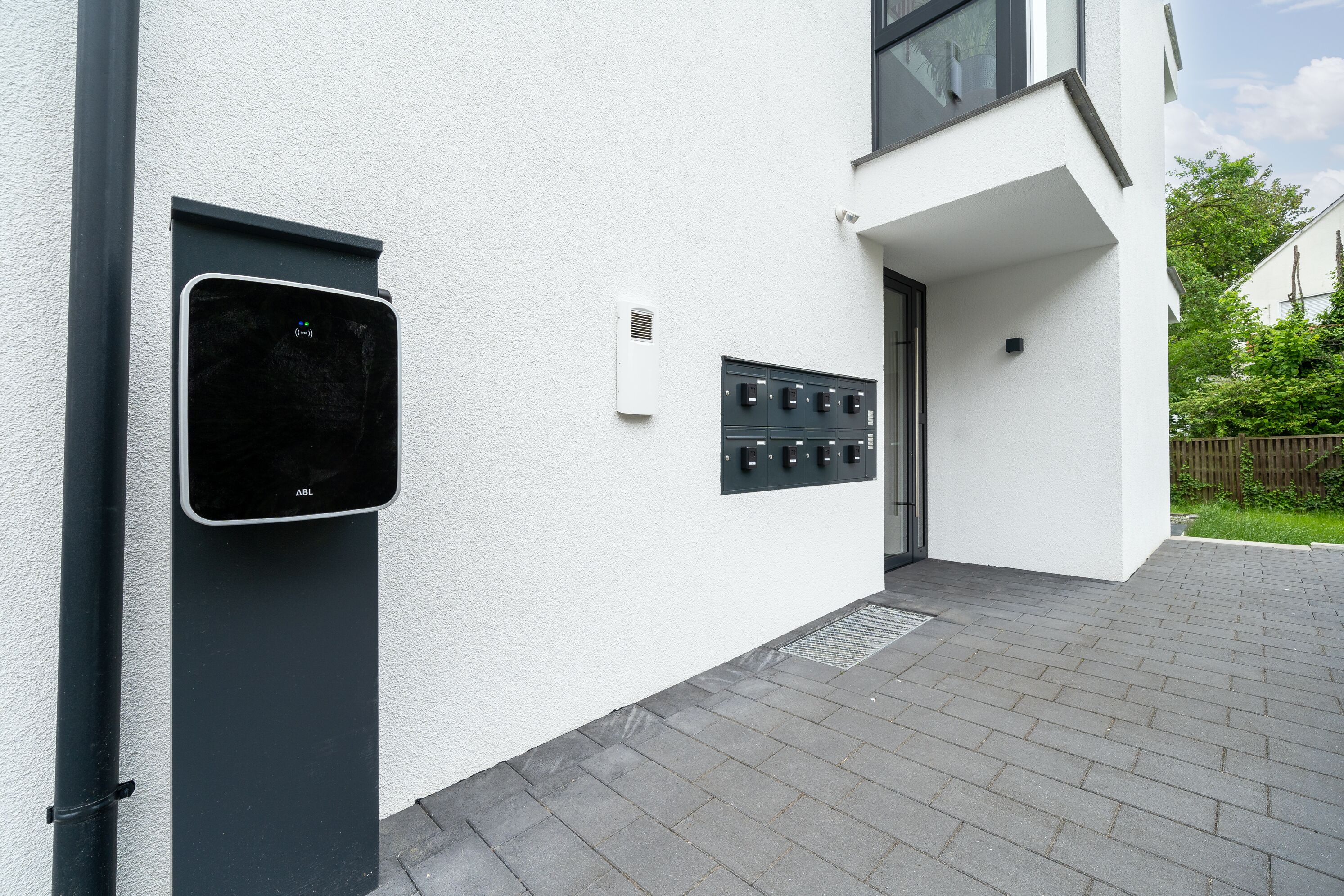 A modern building exterior with a door, a wall-mounted control panel, and a paved entrance area.