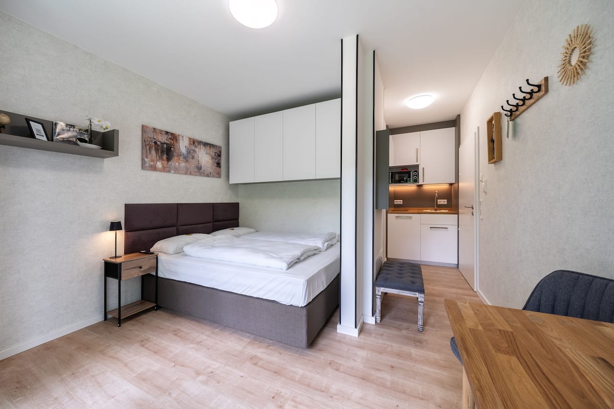 A modern bedroom with a bed, bedside table, and a small desk. There is a kitchenette in the background.
