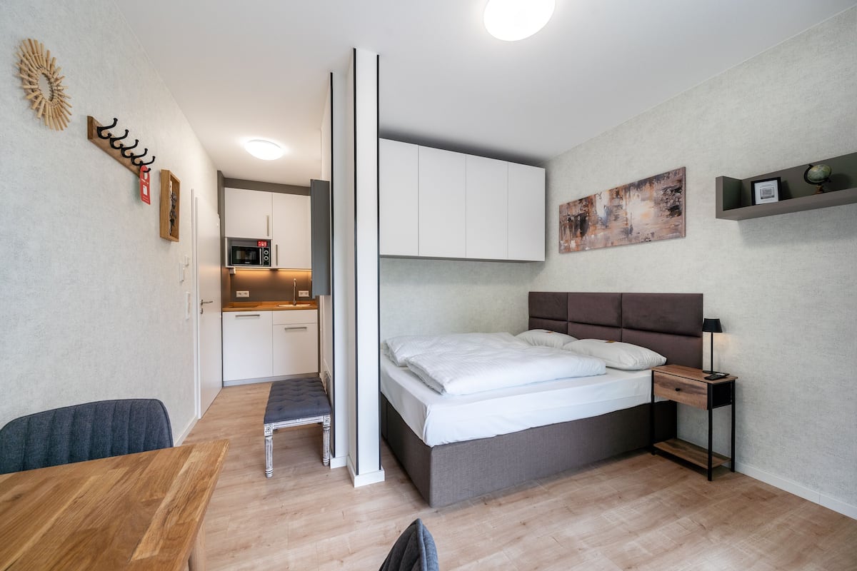 A modern bedroom with a bed, bedside table, and a small desk. There is a kitchenette in the background.