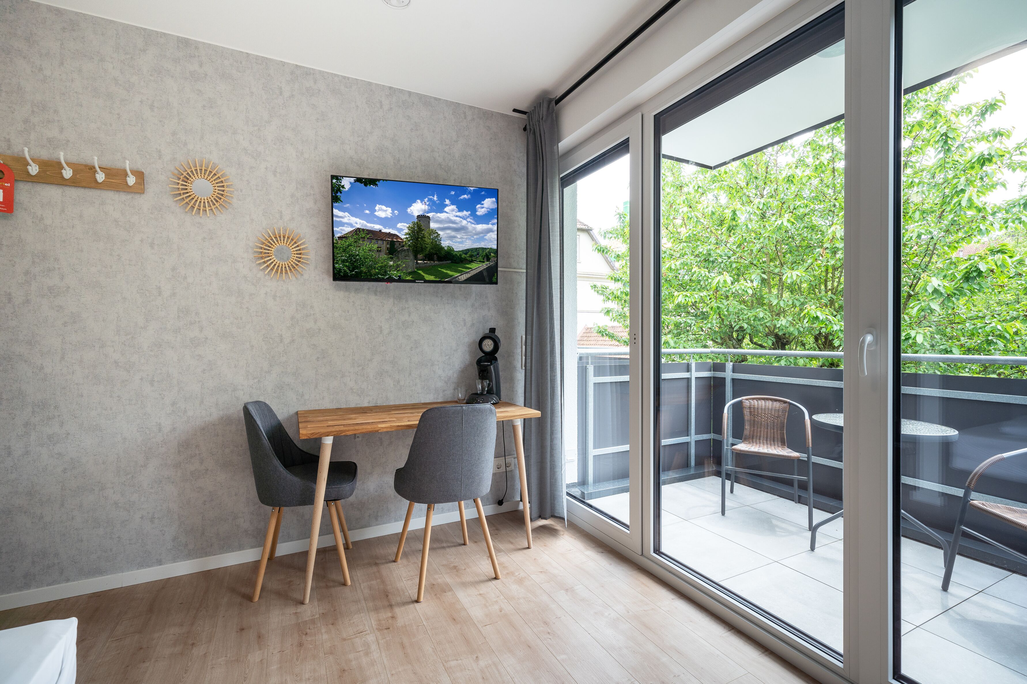 Une chambre moderne avec une table en bois, deux chaises, un téléviseur fixé au mur et un balcon aménagé avec du mobilier d’extérieur.