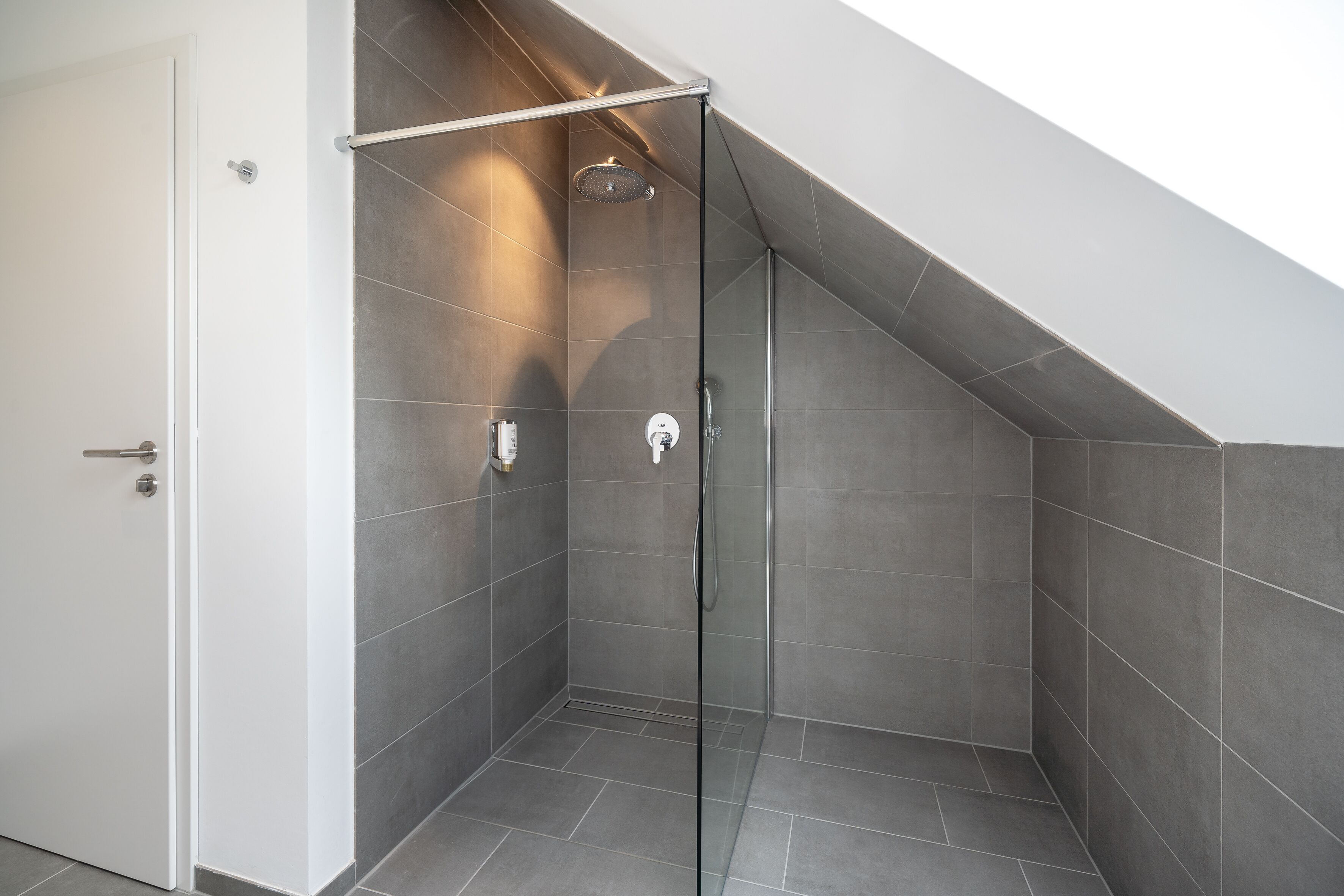Ducha moderna con puerta de cristal, azulejos grises y cabezal de ducha visible.