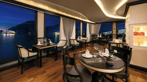A dining area with tables set for four, large windows with curtains, and a view of a body of water at night. -  Le Journey Luxury Cruise (Tuan Chau)