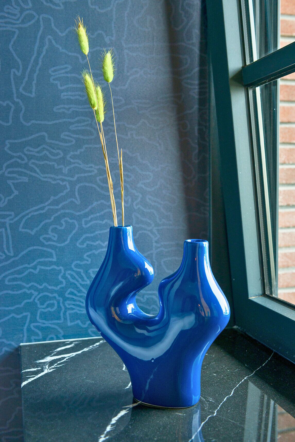 A blue vase with a unique shape on a black surface near a window with a brick wall.