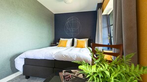 A modern hotel room with a neatly made bed, a small table with a magazine, and a potted plant.