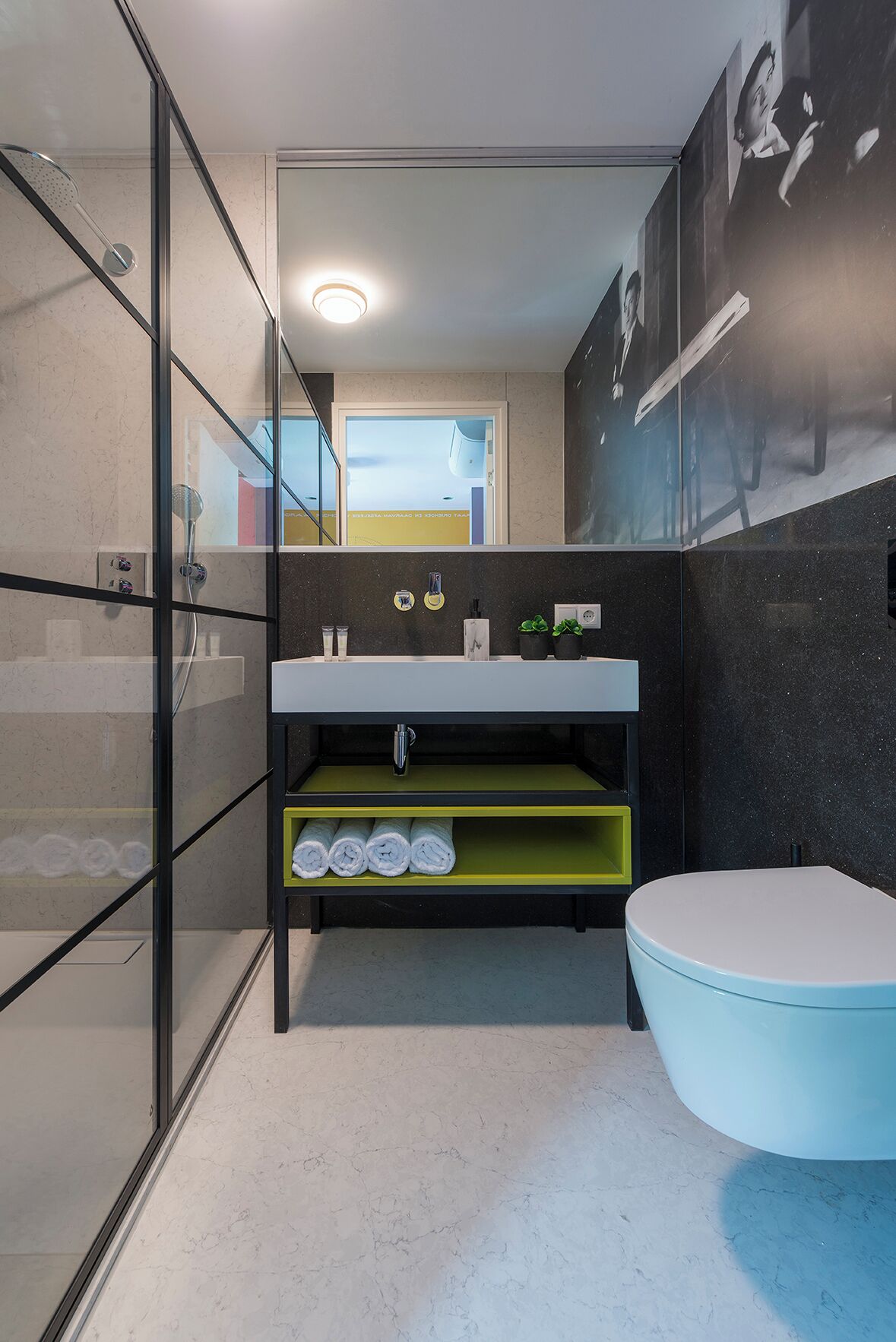 Une salle de bain moderne avec une douche à parois de verre, un lavabo blanc doté d’une étagère verte et des toilettes bleues.