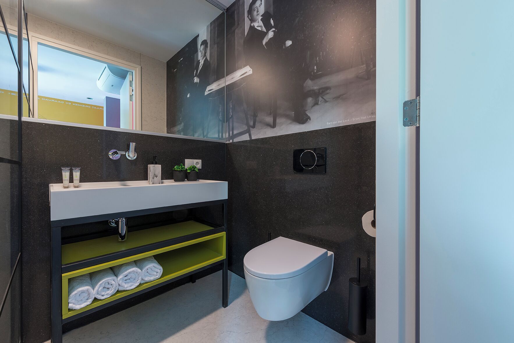 A compact bathroom with a white sink, black and yellow shelves, a toilet, and a wall-mounted mirror.