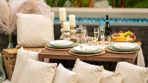 Mesa de comedor dispuesta con platos, vasos y velas, rodeada de almohadones y una cesta, con una piscina y follaje al fondo.