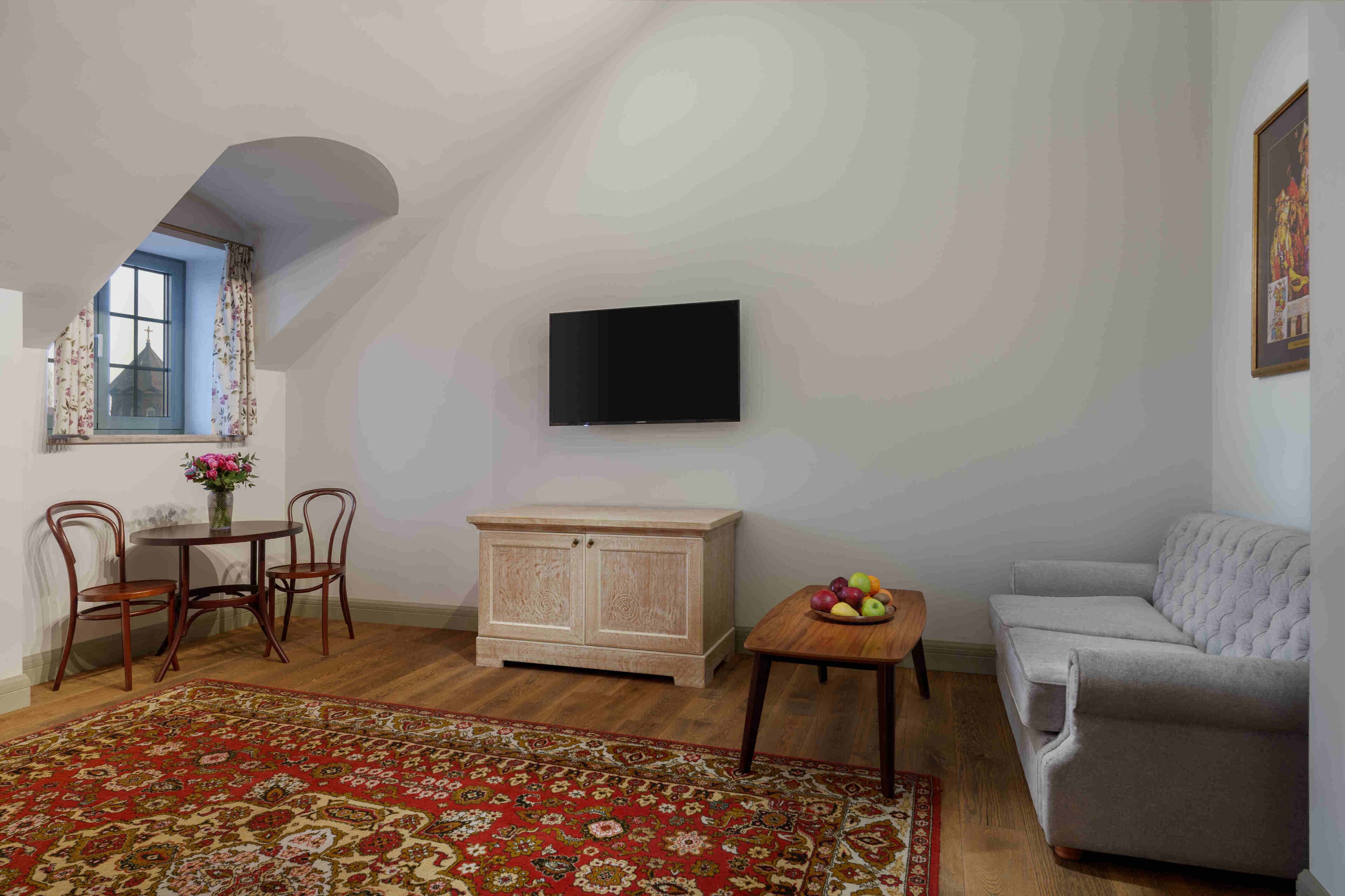 Un salon chaleureux avec un plafond haut, un téléviseur à écran plat fixé au mur, un canapé gris, un meuble en bois, une petite table ronde avec des fruits et un tapis à motifs.