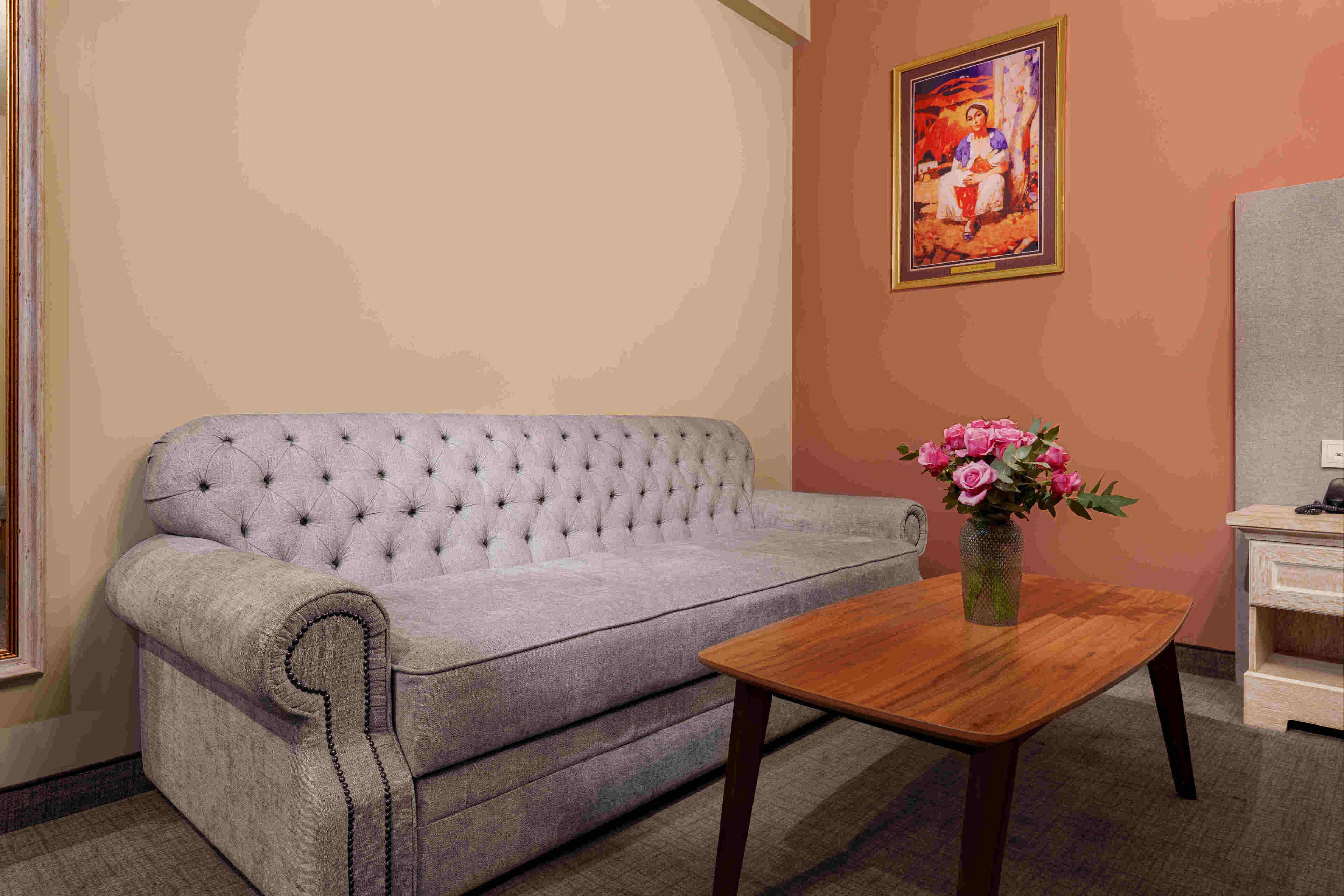A room with a tufted sofa, a wooden table with a vase of flowers, and a painting on the wall.