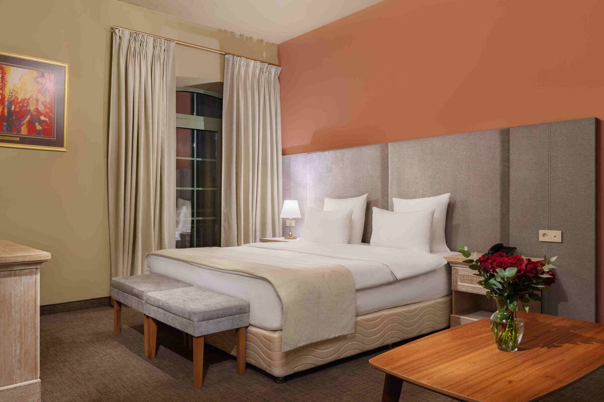 Une chambre d’hôtel avec un lit, une banquette, une table de chevet avec une lampe, un vase de fleurs et un tableau au mur.