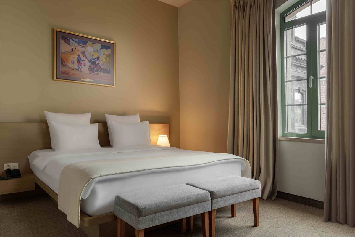 Une chambre d’hôtel avec un grand lit, une table de chevet avec une lampe, une fenêtre avec des rideaux et un tableau au mur.