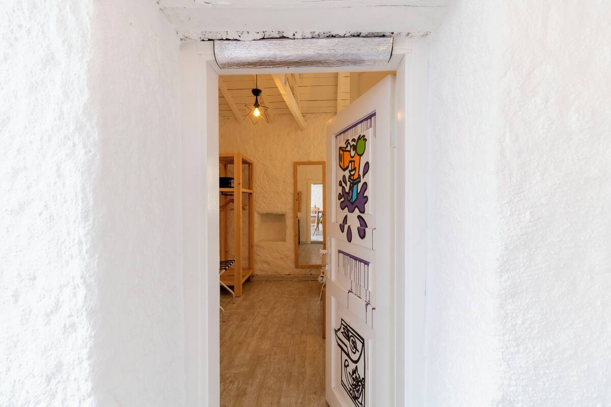 A narrow hallway with white walls and a wooden floor. There is a door on the right with colorful artwork.