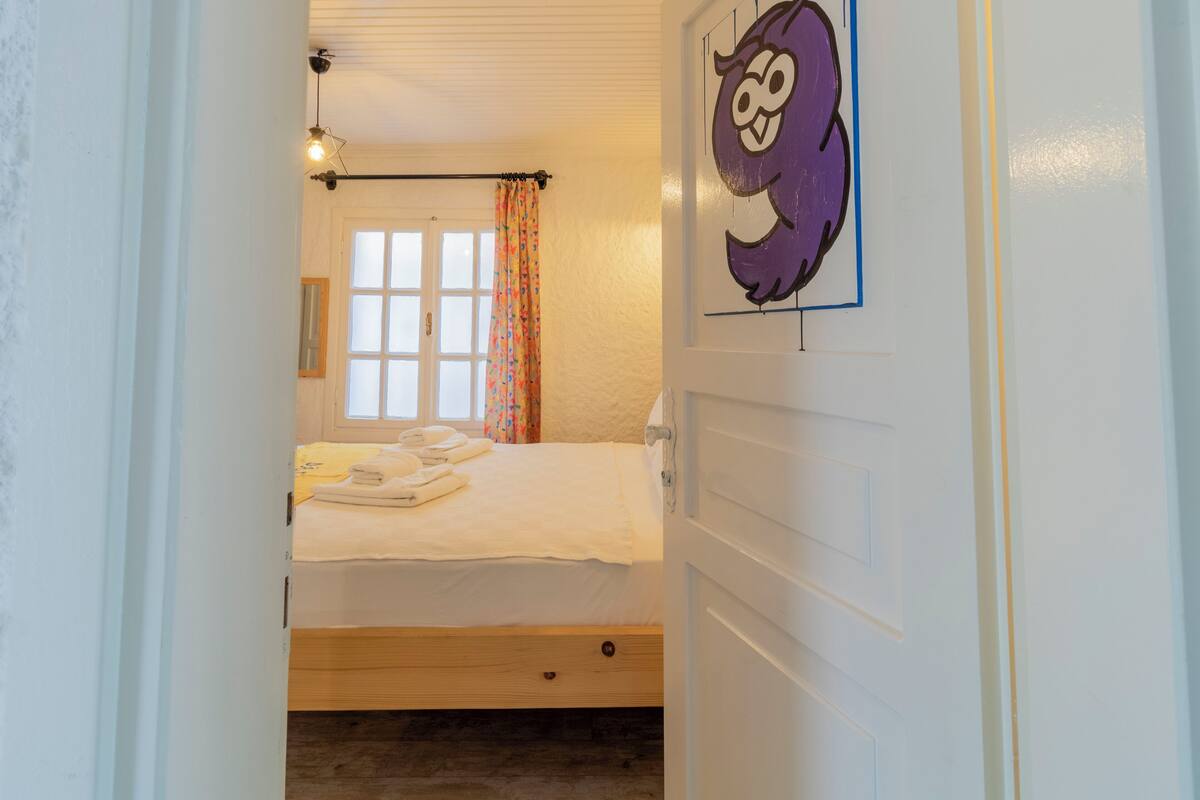 A bedroom with a bed, a window with curtains, a wall-mounted light fixture, and a framed picture.