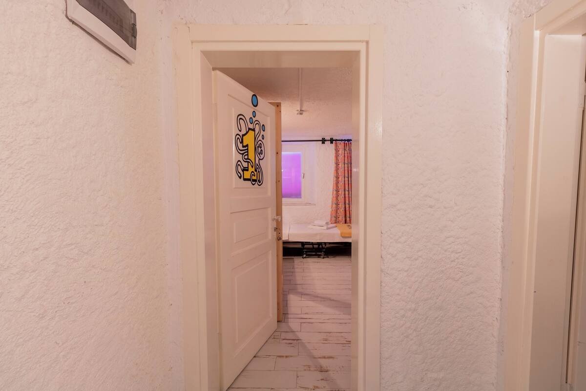 A narrow hallway with a white door on the left, a white door on the right, and a bed with a purple light in the background.