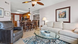 A living room with a glass coffee table, a leather sofa, a white sofa, a dining area with wooden chairs, and a ceiling fan. - Gorgeous oceanfront vacation rental condo with amazing views and pool (North Myrtle Beach)