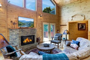 A cozy cabin interior with a stone fireplace, comfortable seating, and a television mounted on the wall. - Scenic Mountain View w/ Hot Tub & Fire Pit (Blue Ridge)