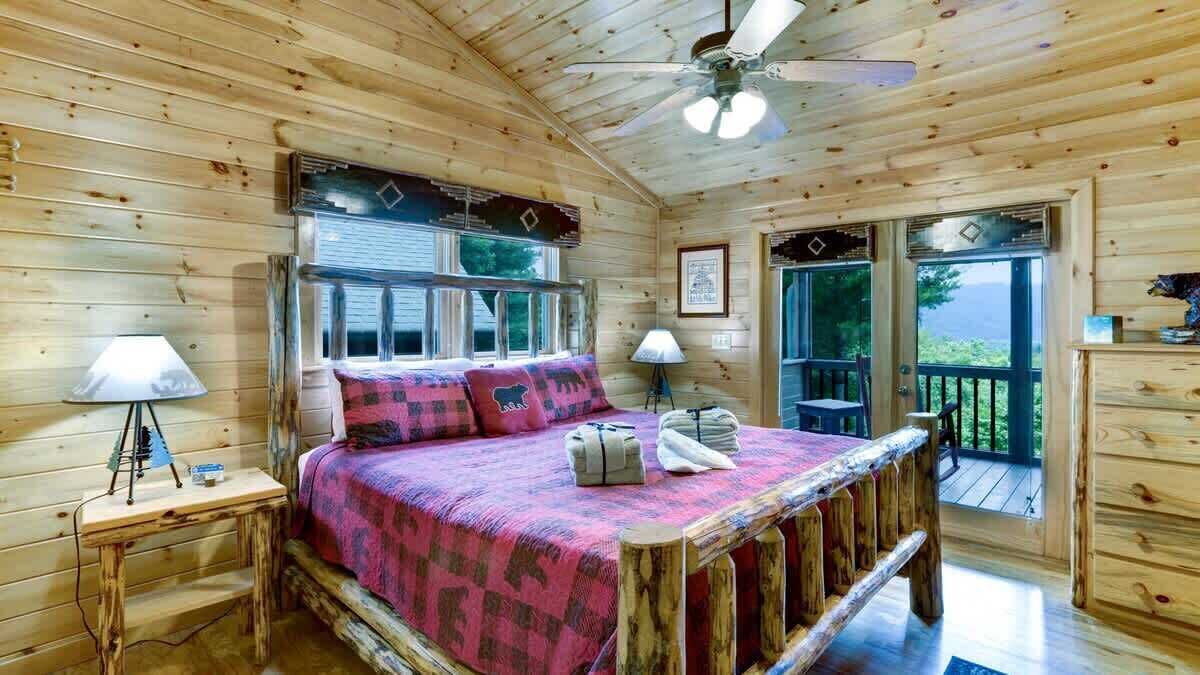 Camera da letto in una baita di legno con un letto, due lampade ai comodini, una ventola a soffitto e vista all'esterno attraverso un'ampia finestra.
