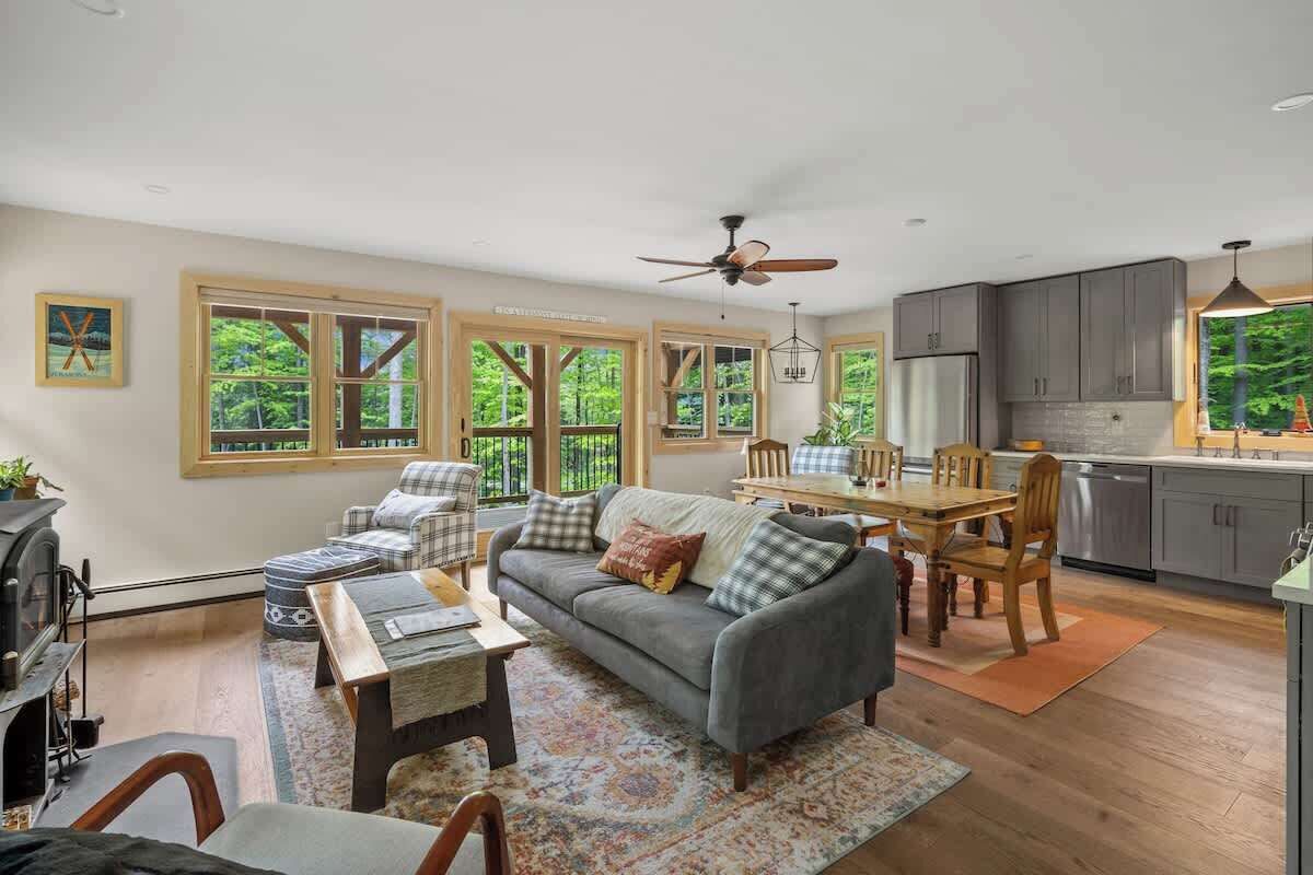 A spacious living room with a gray sofa, wooden dining table, and chairs, a fireplace, and large windows with a view of greenery.