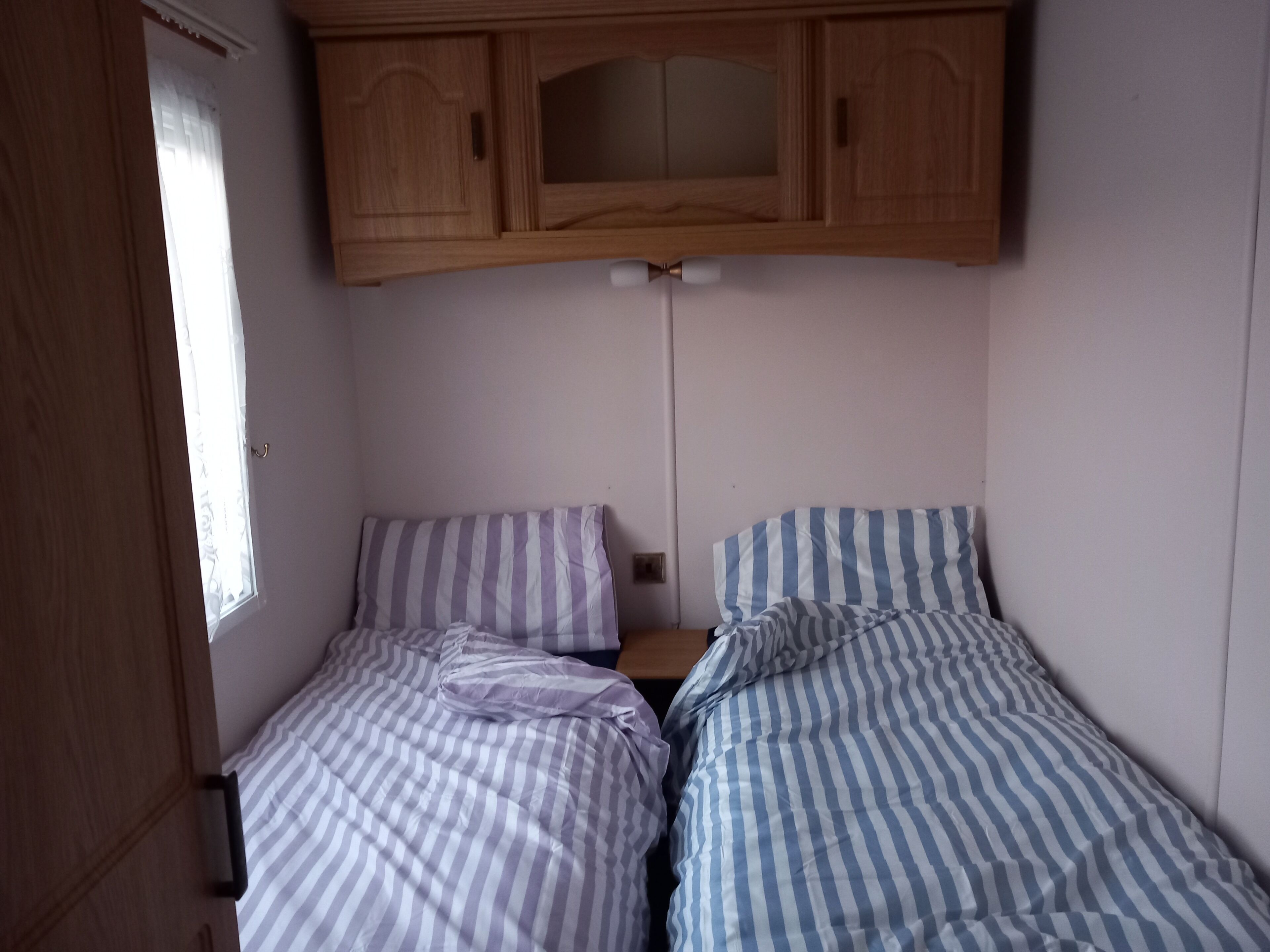 A small, single-room accommodation with two beds, wooden cabinets above, and a window providing natural light.