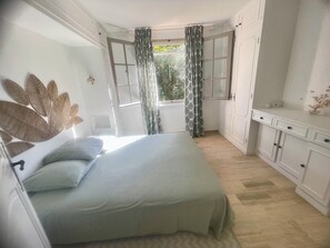 Quarto com cama, janela com cortinas, guarda-roupa e vista para o verde lá fora.