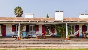 A two-story building with a tiled roof, a covered porch with outdoor seating, and a palm tree in the background. - Nômade Melides Eco Lodge (Grandola)