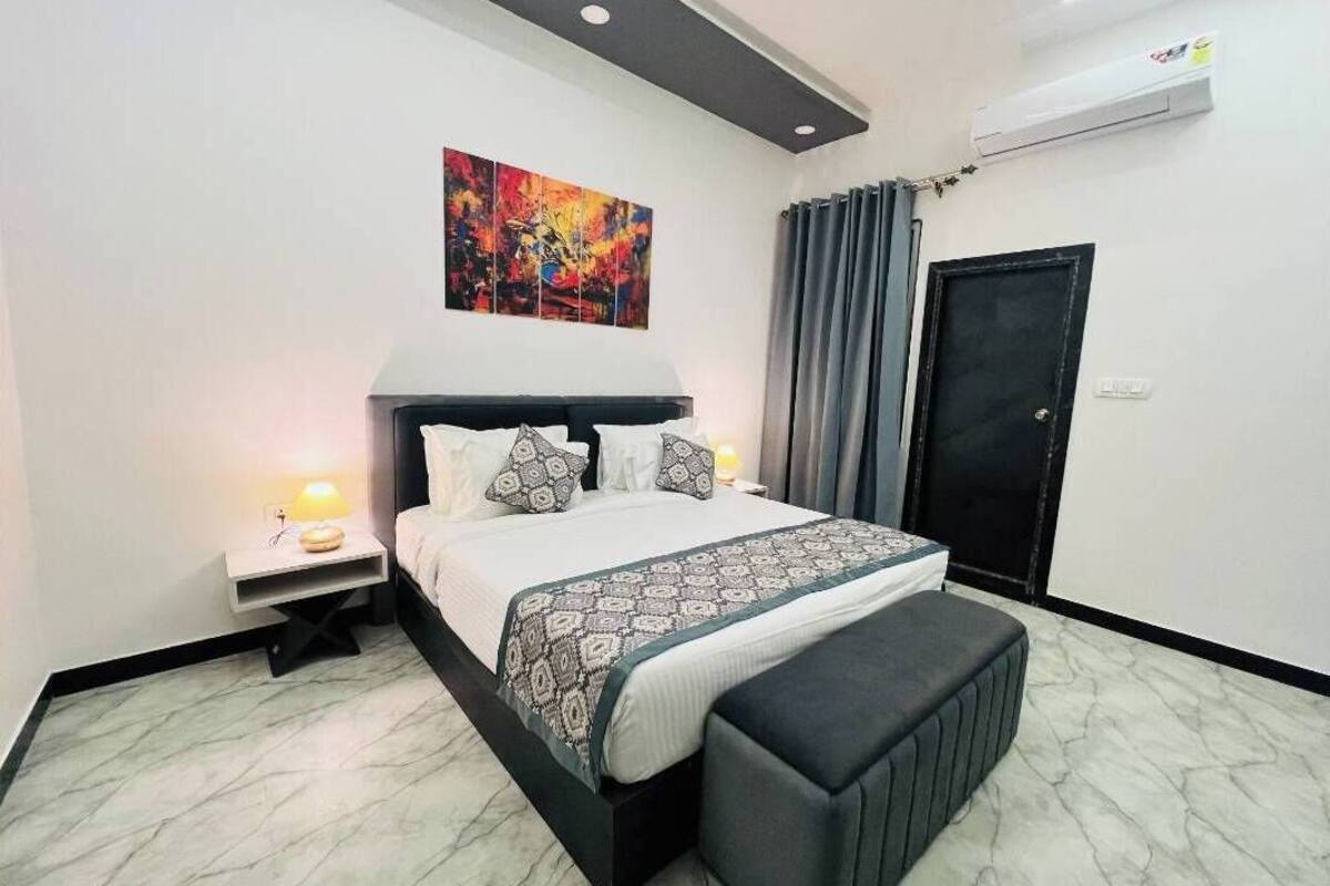 Een moderne slaapkamer met een groot bed, een nachtkastje met een lamp, een bankje aan het voeteneind van het bed, een kleurrijk abstract schilderij boven het bed en een deur met gordijn.