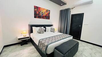 A modern bedroom with a large bed, a bedside table with a lamp, a bench at the foot of the bed, a colorful abstract painting above the bed, and a door with a curtain.