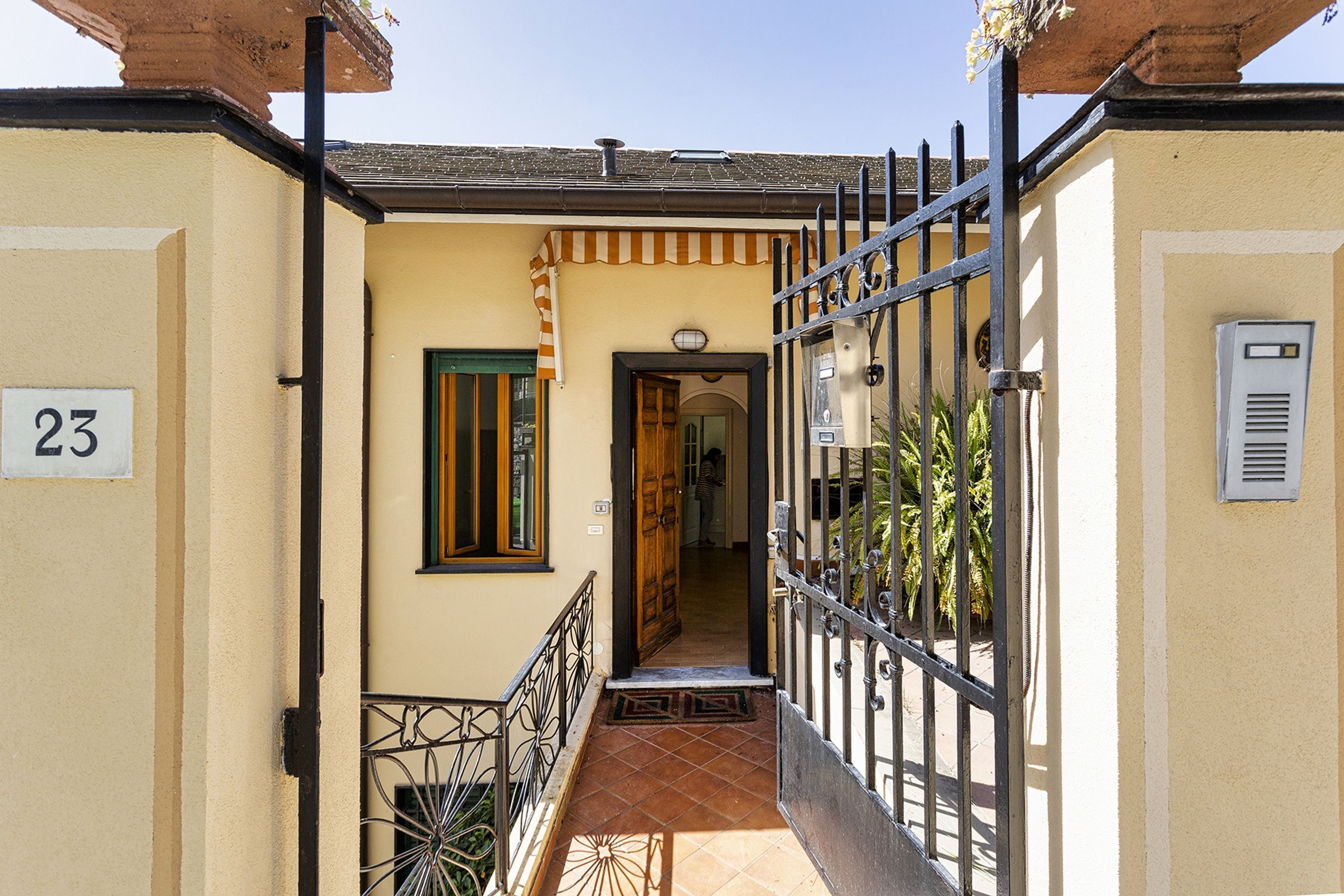 A narrow hallway with a tiled floor leading to a door with a striped awning. A number '23' is visible on the wall to the left.