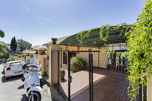 Une allée privée fermée par un portail, où sont garés un scooter et une voiture, mène à un bâtiment doté d’un toit en tuiles et d’une pergola couverte de plantes grimpantes.