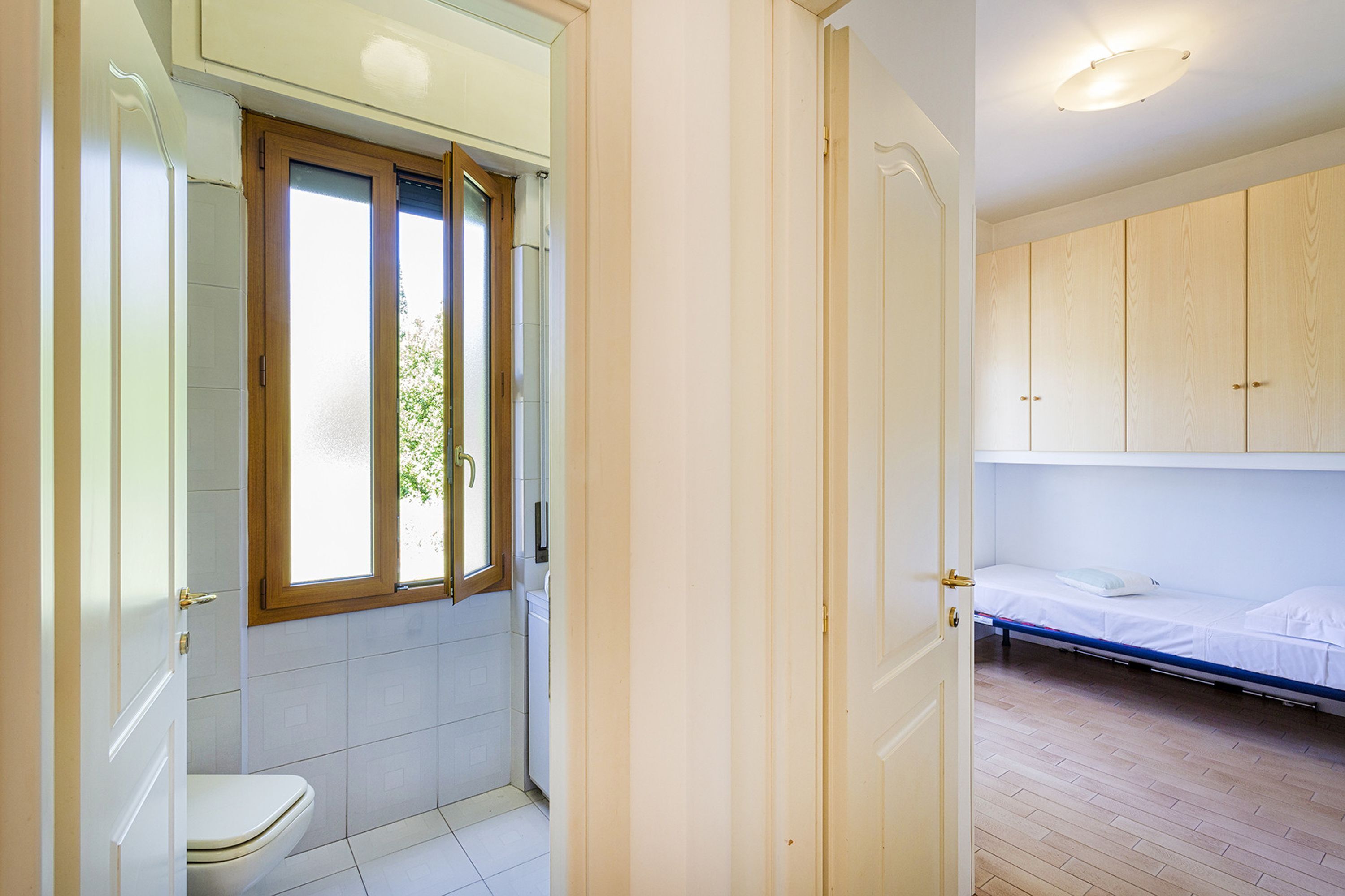 Een badkamer met toilet, een raam en een douche. Er is een deur naar een slaapkamer met een bed en houten kasten.