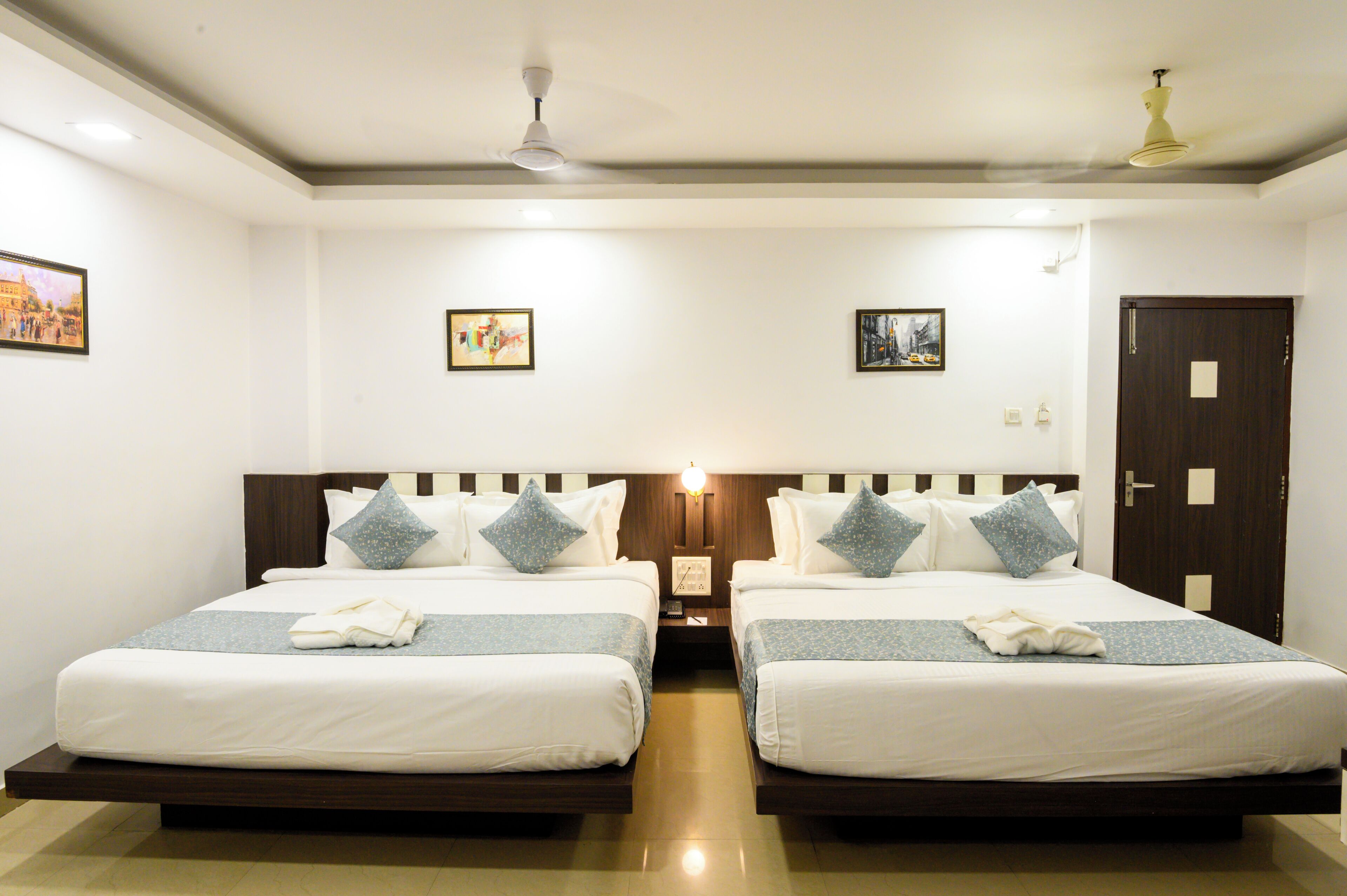 Quarto de hotel com duas camas de solteiro, cabeceira de madeira, mesa de cabeceira com abajur e porta com fechadura.