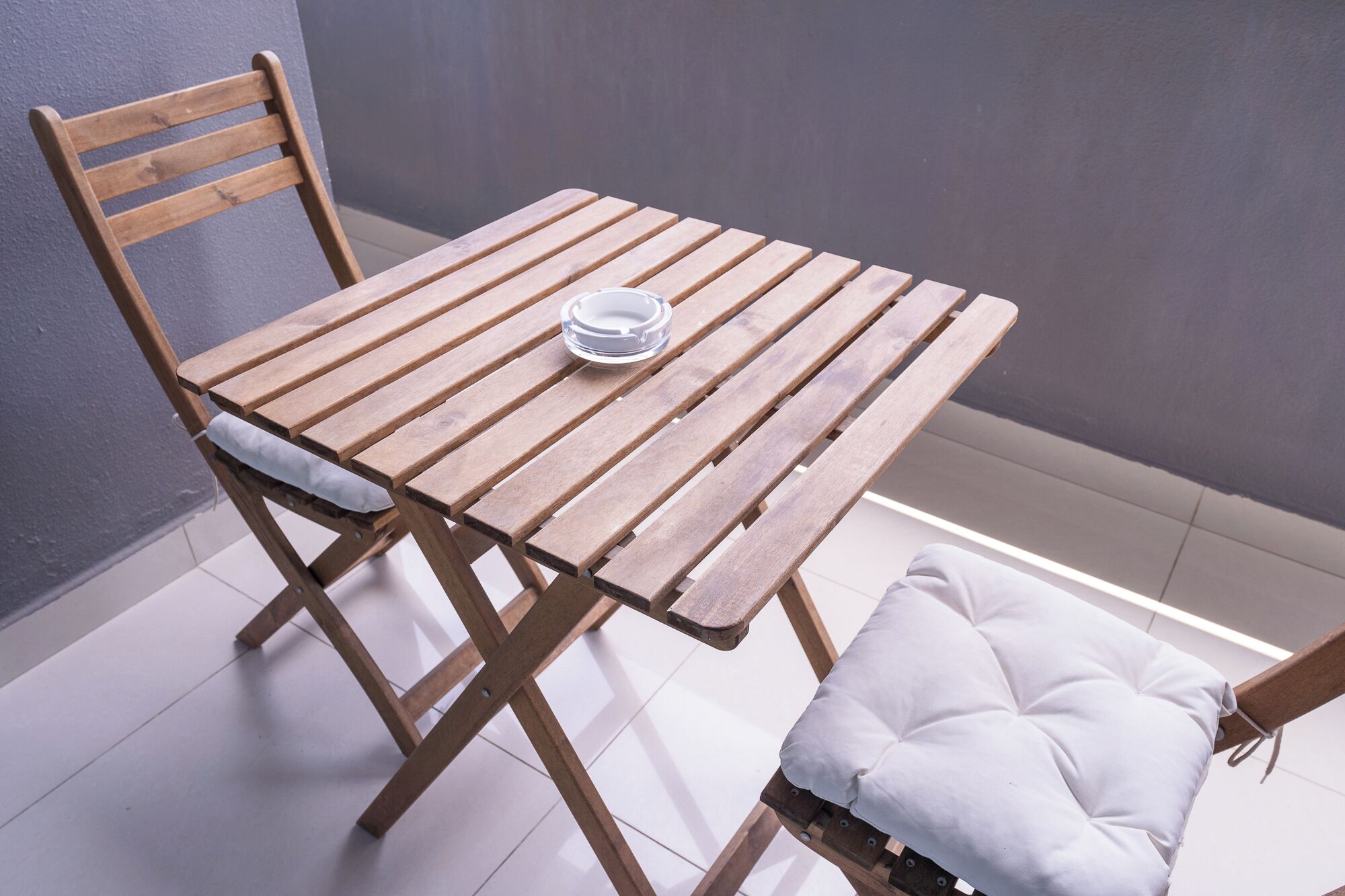 Een houten tafel met een glazen schaal, een fauteuil met kussen, en een wit kussen op een betegelde vloer tegen een grijze muur.