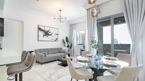 Een moderne woonkamer met een grijze bank, een glazen eettafel voor vier personen en een groot raam met gordijnen.