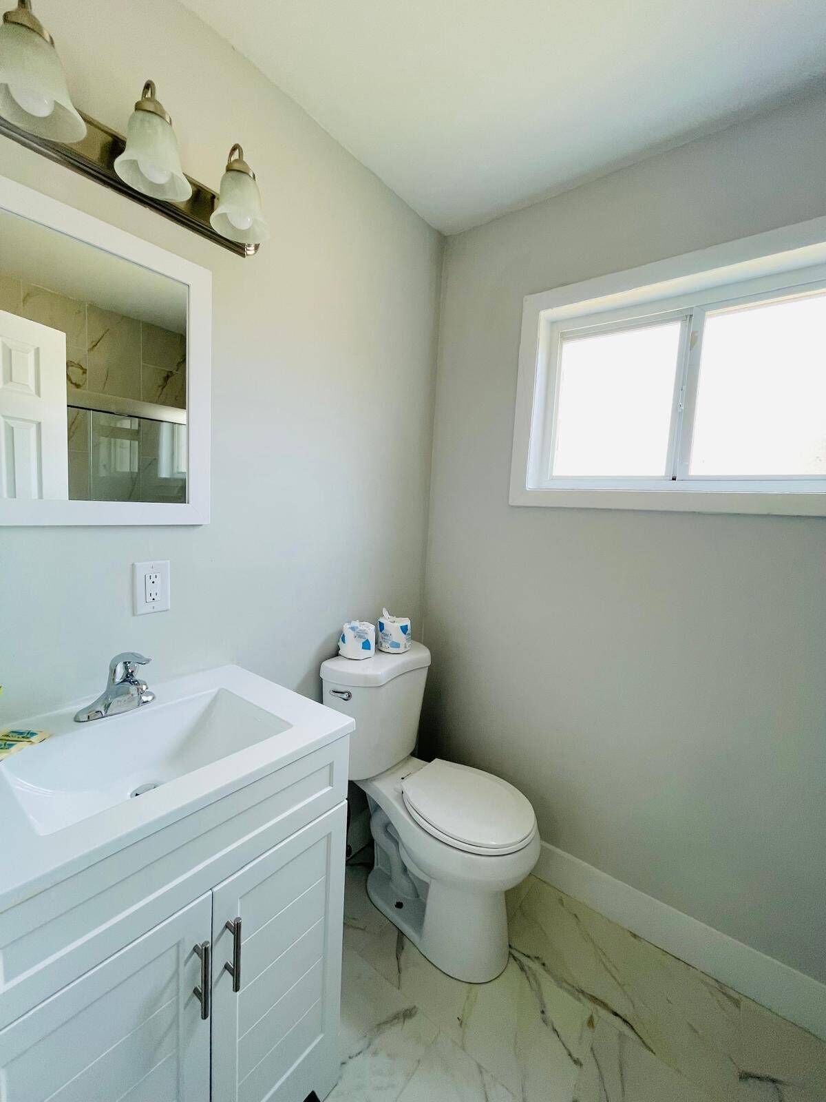 Un baño con un lavabo blanco, espejo e inodoro. Hay una ventana sobre el inodoro y una lámpara sobre el espejo.
