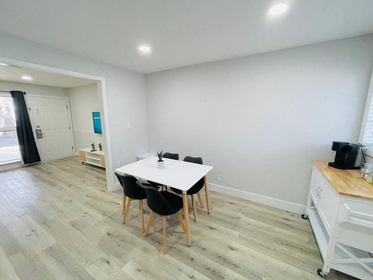 Quarto moderno e bem iluminado com uma mesa e cadeiras brancas, uma pequena planta e uma máquina de café sobre um balcão de madeira.