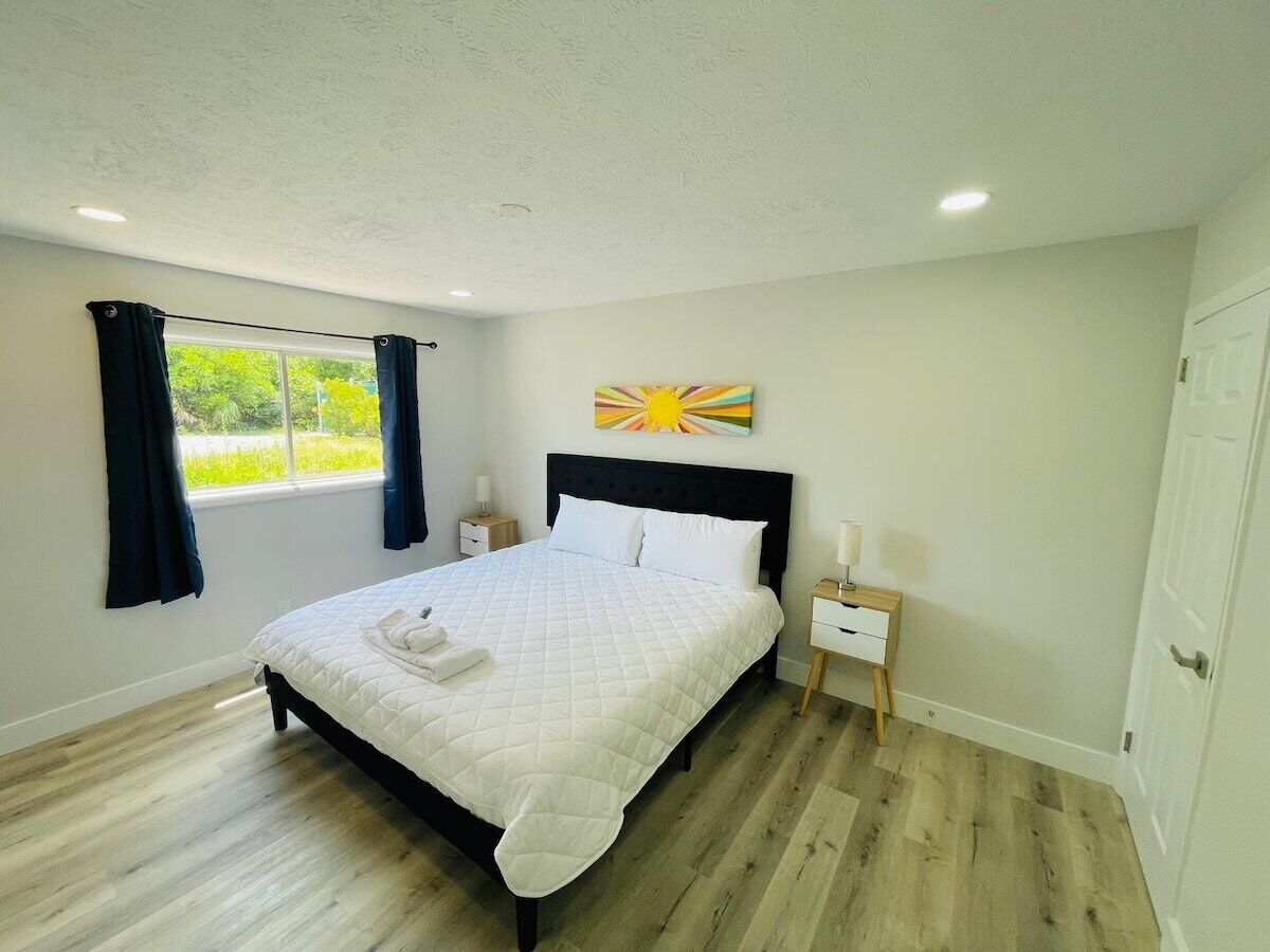 A bedroom with a bed, a nightstand, a window with curtains, a wall art piece, and a door.