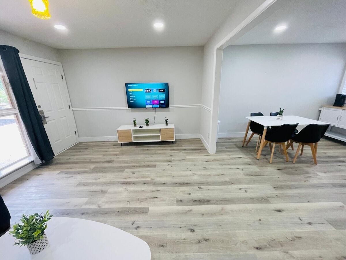 A modern living room with a flat-screen TV mounted on the wall, a white entertainment unit, a dining area with a table and chairs, and a wooden floor. - Perfect vacation walk to the beach 6 (Myrtle Beach)