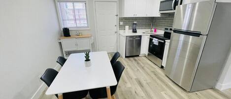 A compact kitchen with white cabinets, a stainless steel refrigerator, a microwave, and a dining area with a white table and black chairs.
