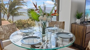 A dining table set with clear glassware and plates, surrounded by wicker chairs, with a view of palm trees and the ocean.