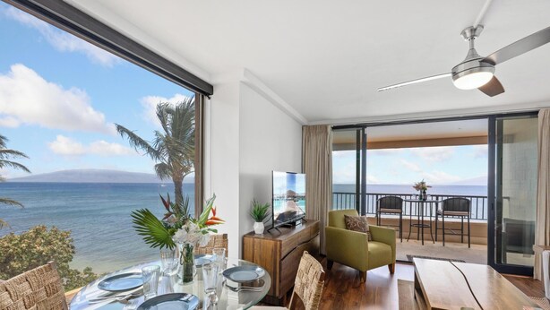A modern living room with a glass dining table, comfortable seating, a flat-screen TV, and a balcony with a view of the ocean.