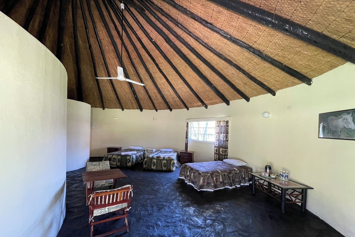 A room with a high ceiling, a ceiling fan, and a bed with a patterned bedspread. There is a small table with a lamp and a chair.