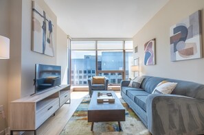 Una sala de estar moderna con un televisor de pantalla plana, un sofá gris, una mesa de centro y un ventanal con vistas a la ciudad.