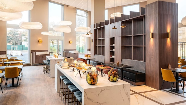 A modern restaurant interior with a central bar, seating areas, and large windows.
